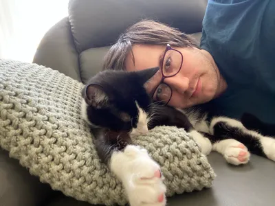 Person resting beside a tuxedo cat and knitted cushion